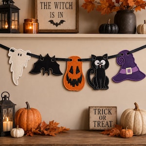 May include: A Halloween garland featuring five felt cutouts strung on a black ribbon: a white ghost, a black bat, an orange jack-o'-lantern, a black cat with large white eyes, and a purple witch hat with a light purple band and buckle.