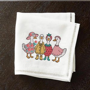 K&ouml;nnte beinhalten: Besticktes K&uuml;chentuch mit vier G&auml;nsen, die verschiedene Outfits tragen. Die G&auml;nse tragen einen rosa Hut, einen gelben Regenmantel, ein Erdbeerkost&uuml;m und ein rosa Kleid. Das Handtuch hat einen wei&szlig;en Hintergrund mit einer Spitzenborte.