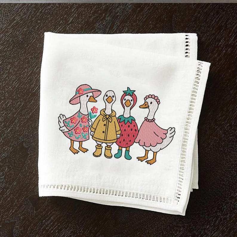 K&ouml;nnte beinhalten: Besticktes K&uuml;chentuch mit vier G&auml;nsen, die verschiedene Outfits tragen. Die G&auml;nse tragen einen rosa Hut, einen gelben Regenmantel, ein Erdbeerkost&uuml;m und ein rosa Kleid. Das Handtuch hat einen wei&szlig;en Hintergrund mit einer Spitzenborte.