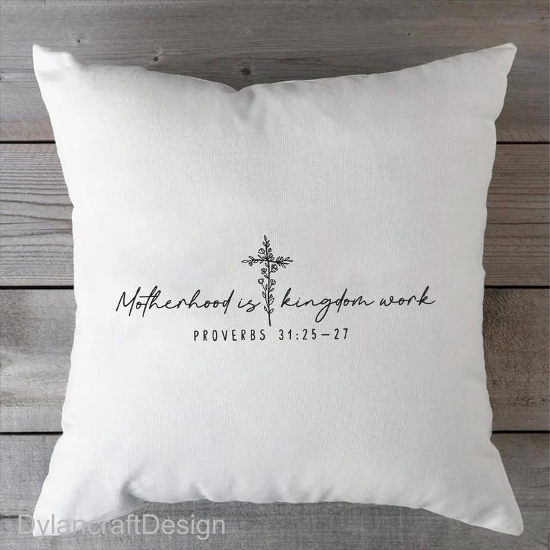 Puede incluir: Coj&iacute;n cuadrado blanco con el texto "Motherhood is kingdom work" y un dise&ntilde;o de cruz floral. El texto "PROVERBIOS 31:25-27" est&aacute; debajo. El coj&iacute;n est&aacute; sobre una superficie de madera.