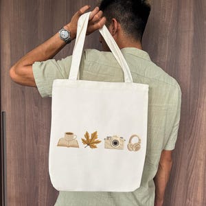 May include: Cream-colored tote bag with embroidered images of a coffee cup, open book, maple leaf, camera, and headphones. The bag has two long handles and is held by a person.