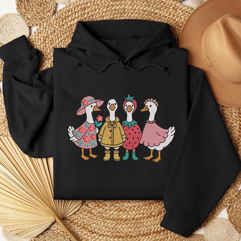 K&ouml;nnte beinhalten: Schwarzer Kapuzenpullover mit einem Aufdruck von vier G&auml;nsen, die verschiedene Outfits tragen. Die G&auml;nse tragen einen rosa Hut, einen gelben Regenmantel, ein Erdbeerkost&uuml;m und ein rosa Kleid.