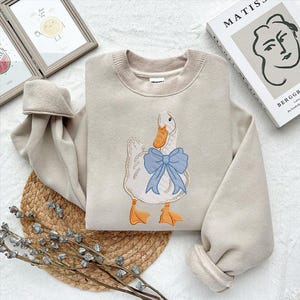 May include: A beige sweatshirt with a white duck embroidered on the front. The duck has a blue bow around its neck.