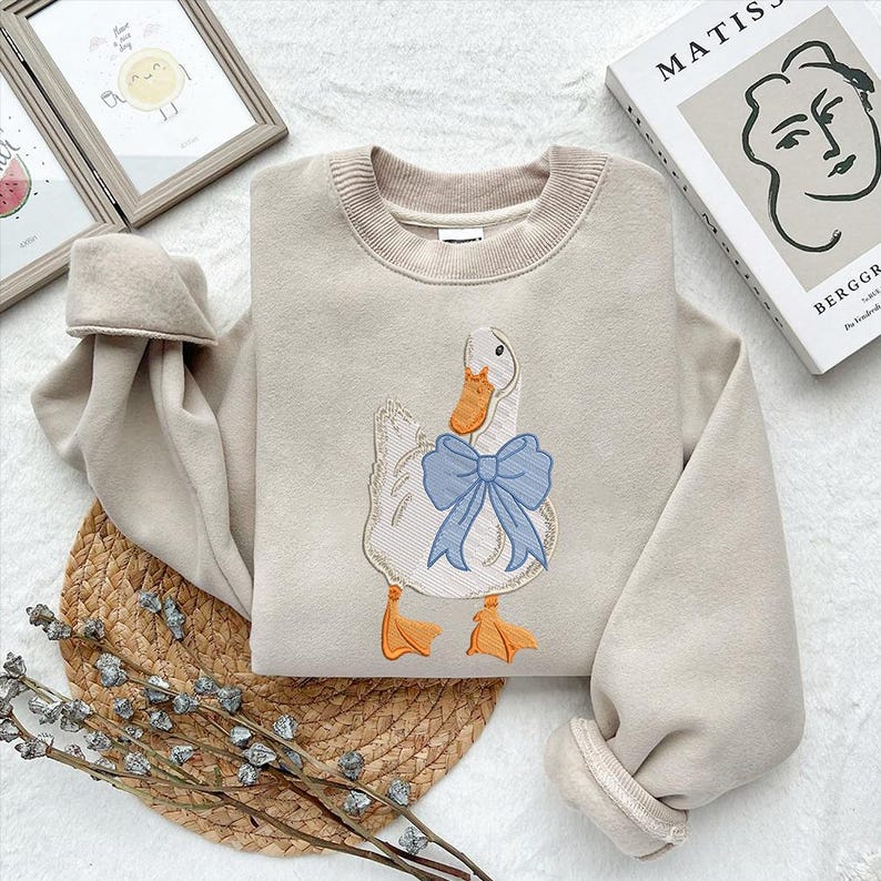May include: A beige sweatshirt with a white duck embroidered on the front. The duck has a blue bow around its neck.