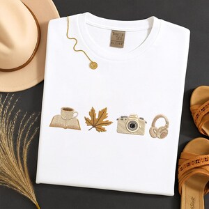 May include: White crew neck t-shirt with embroidered fall-themed images: a coffee cup, open book, maple leaf, camera, and headphones. A gold necklace and a beige hat are also visible.