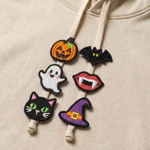 Puede incluir: Una sudadera con capucha de color beige con dos cordones de color crema decorados con parches bordados de Halloween. Los parches incluyen una calabaza naranja, un fantasma blanco, un gato negro, un murciélago, labios rojos y un sombrero de bruja.