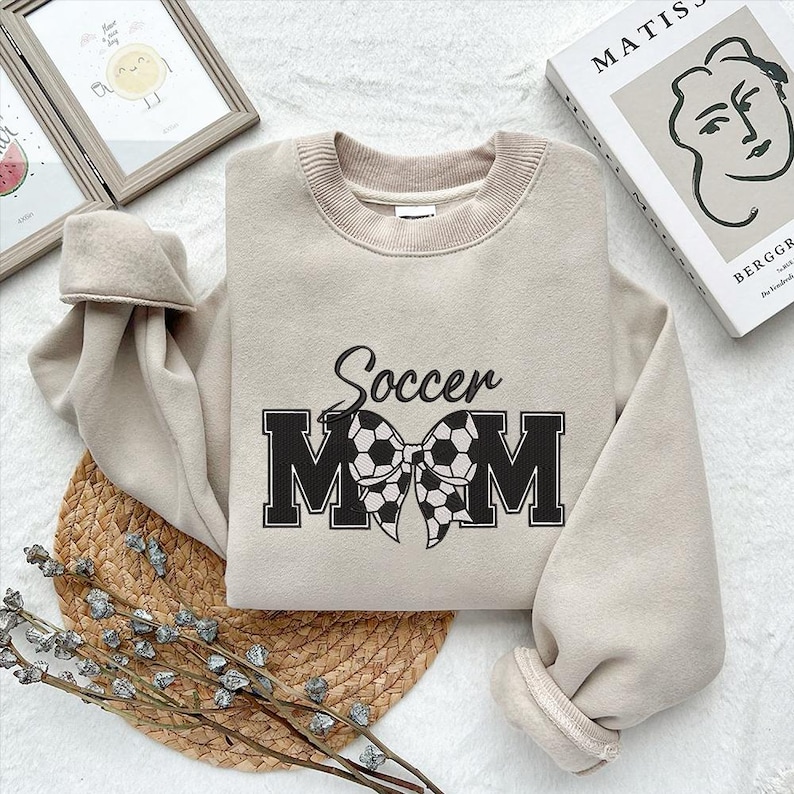 Peut inclure: Un sweat-shirt beige avec le texte "Soccer Mom" en noir avec un n&oelig;ud de ballon de football noir et blanc.