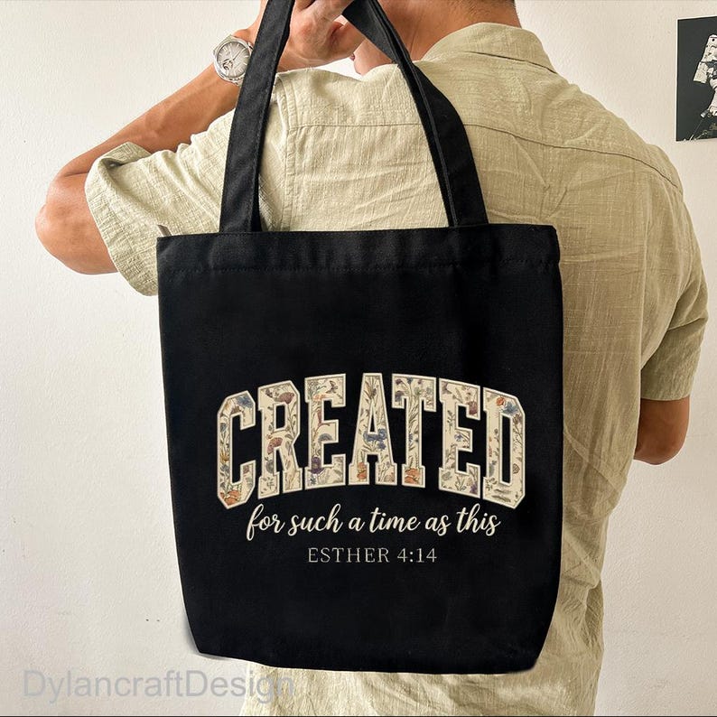 May include: Black tote bag featuring the word "CREATED" in a floral design, with the text "for such a time as this" and "ESTHER 4:14" below. The bag has black handles and is being held by a person.