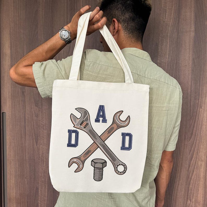 May include: Cream-colored tote bag featuring a design with crossed wrenches, a bolt, and the word "DAD" in blue block letters. The bag has two cream-colored handles.