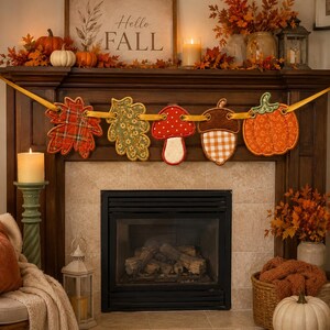 May include: A fabric garland featuring five hanging ornaments strung on a yellow ribbon: a red plaid leaf, a green floral leaf, a red mushroom with white spots, a brown acorn with a checkered pattern, and an orange pumpkin with a green stem.