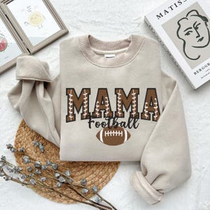 May include: A beige sweatshirt with the text "MAMA Football" in brown letters with stitching detail. A brown football graphic is below the text.