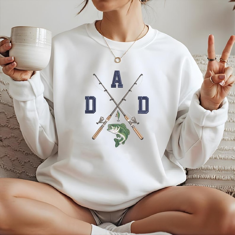 May include: White sweatshirt with a graphic design featuring the word "DAD" in navy blue block letters, crossed fishing rods, and a green fish. The sweatshirt is long-sleeved and has a crew neck.