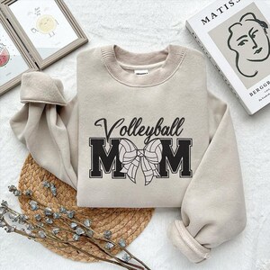 Peut inclure: Un sweat-shirt beige avec le texte "Volleyball Mom" en lettres noires avec un graphique de volley-ball noir et blanc et un n&oelig;ud.