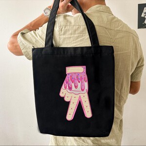 May include: Black tote bag with a pink and white hand with sprinkles and icing design. The hand is making a peace sign.