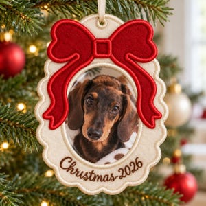 May include: An oval-shaped felt Christmas ornament featuring a brown and black dachshund photograph in the centre. The ornament has a scalloped cream-coloured border, a large red embroidered bow at the top, and the text Christmas 2026 in brown.