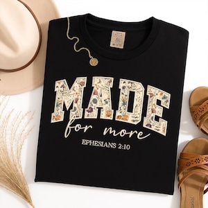 Può includere: T-shirt nera con la scritta "MADE for more" in un design floreale. Il testo "Ephesians 2:10" è in basso. Sono visibili anche una collana d'oro e sandali marroni.
