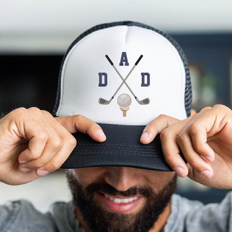 May include: White and black trucker hat with the word "DAD" in navy blue block letters, crossed golf clubs, and a golf ball on a tee. The hat has a mesh back and a curved brim.