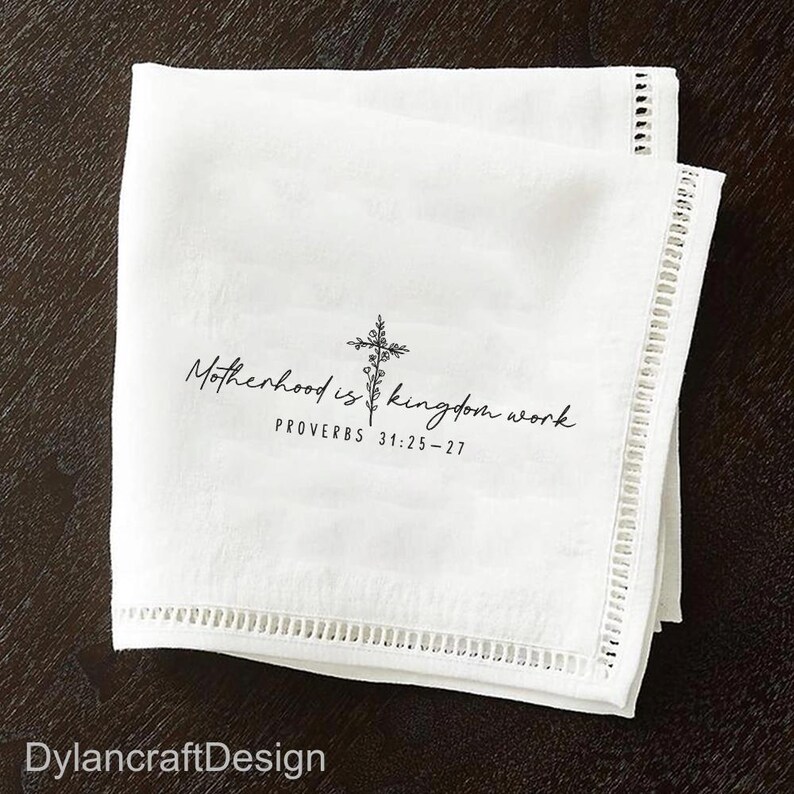 Puede incluir: Servilleta de lino blanco con un borde calado decorativo. La servilleta presenta el texto "Motherhood is kingdom work" con un dise&ntilde;o de cruz y floral, y el vers&iacute;culo b&iacute;blico Proverbios 31:25-27.