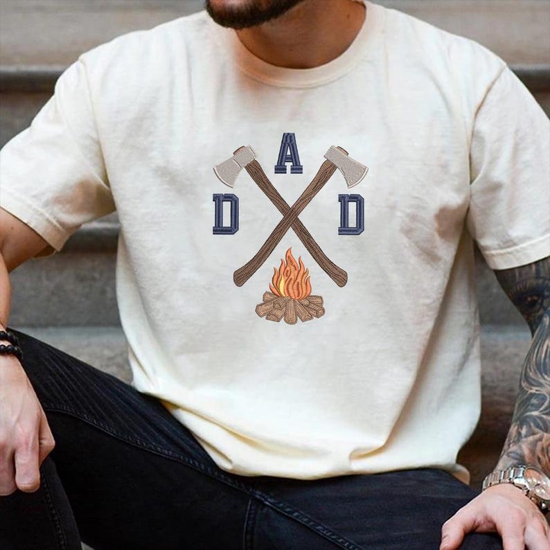 May include: Cream-colored t-shirt featuring a graphic design with the word "DAD" and crossed axes. The design includes a campfire illustration. The shirt is short-sleeved and has a crew neck.