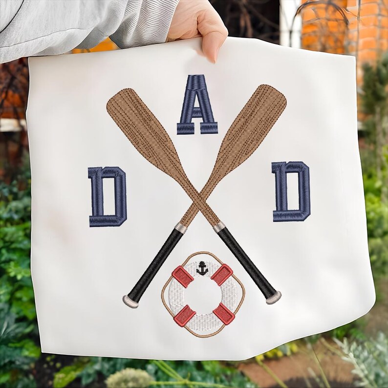 May include: White fabric with an embroidered design featuring the word "DAD" in navy blue letters. Two crossed oars in brown are centered, with a red and white life preserver below. The design has a nautical theme.