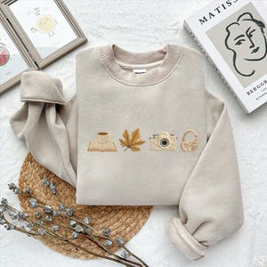 May include: Beige sweatshirt with embroidered fall-themed icons: a book with a coffee cup, a maple leaf, a camera, and headphones. The sweatshirt has a ribbed crew neck and cuffs. The image also shows a book, a framed picture, and a woven mat.