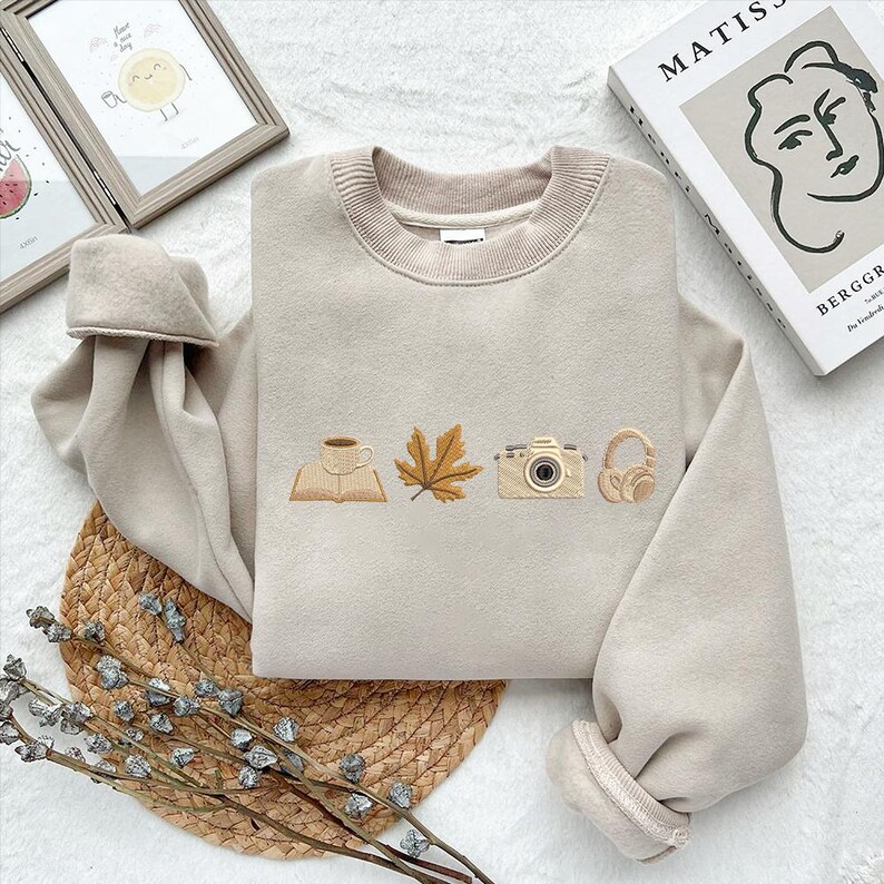May include: Beige sweatshirt with embroidered fall-themed icons: a book with a coffee cup, a maple leaf, a camera, and headphones. The sweatshirt has a ribbed crew neck and cuffs. The image also shows a book, a framed picture, and a woven mat.