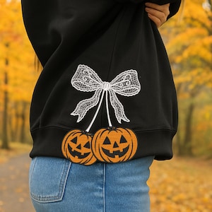 Puede incluir: Sudadera negra con un lazo de encaje blanco y dos calabazas naranjas. Las calabazas tienen caras talladas. La sudadera se lleva sobre unos vaqueros azules, con un fondo otoñal borroso.