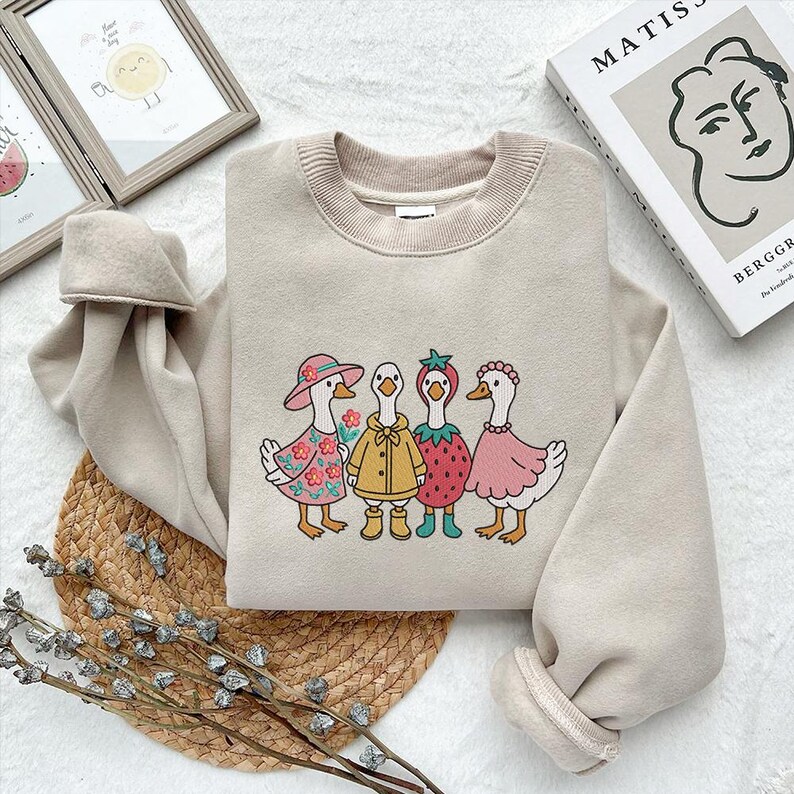 K&ouml;nnte beinhalten: Ein beiger Pullover mit einem Aufdruck von vier G&auml;nsen, die H&uuml;te und Kleider tragen. Die G&auml;nse tragen eine Blumenkrone, einen gelben Regenmantel, einen Erdbeerhut und ein rosa Kleid.