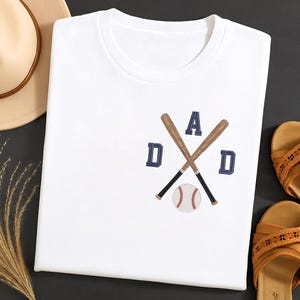 May include: White t-shirt with an embroidered baseball-themed design. The design features crossed baseball bats, the word "DAD" in navy blue letters, and a baseball. The shirt is folded and displayed with sandals and a hat.