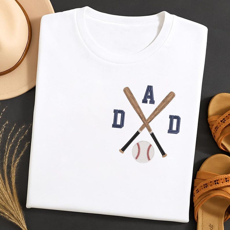 May include: White t-shirt with an embroidered baseball-themed design. The design features crossed baseball bats, the word "DAD" in navy blue letters, and a baseball. The shirt is folded and displayed with sandals and a hat.