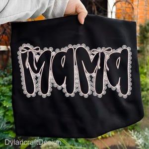 May include: Black tote bag with the word "MAMA" in a white, cursive font, outlined with a delicate lace design. The word "MAMA" has a bow on top of the letters "M" and "A". The tote bag is held by a person.