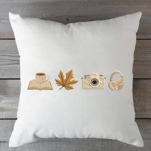 May include: White square pillow with embroidered images of a coffee cup on a book, a maple leaf, a camera, and headphones. The embroidery is in shades of brown and tan.