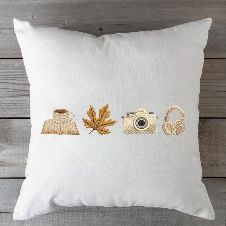 May include: White square pillow with embroidered images of a coffee cup on a book, a maple leaf, a camera, and headphones. The embroidery is in shades of brown and tan.