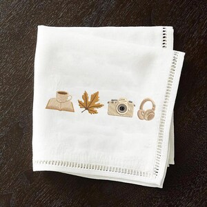 May include: White linen napkin with embroidered images of a coffee cup on a book, a maple leaf, a camera, and headphones. The napkin has a decorative, open-weave border. The embroidered images are in shades of brown and tan.