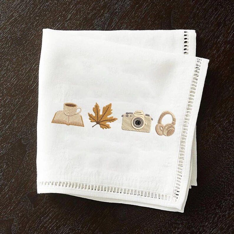May include: White linen napkin with embroidered images of a coffee cup on a book, a maple leaf, a camera, and headphones. The napkin has a decorative, open-weave border. The embroidered images are in shades of brown and tan.