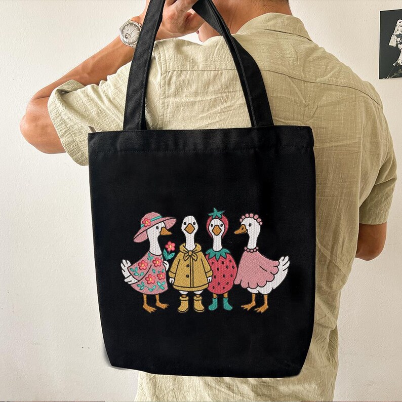 K&ouml;nnte beinhalten: Schwarze Tragetasche mit wei&szlig;em Hintergrund und vier gestickten G&auml;nsen, die verschiedene Outfits tragen. Die G&auml;nse tragen ein rosa Blumenkleid, einen gelben Regenmantel, ein rotes Erdbeerkleid und ein rosa Kleid.