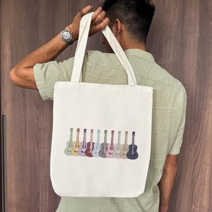May include: Off-white tote bag featuring a row of colorful embroidered guitars. The guitars vary in color, including green, yellow, purple, and blue. The bag has two long handles and is held by a person.