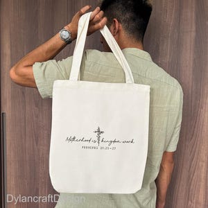 Puede incluir: Bolso tote color crema con el texto "Motherhood is kingdom work" y una cruz. El bolso tiene asas largas y lo sostiene una persona con una camisa verde claro. El texto "PROVERBS 31:25-27" est&aacute; debajo.