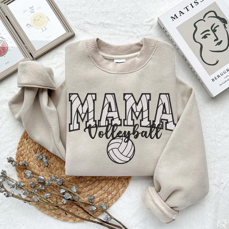 May include: A beige sweatshirt with the text "MAMA Volleyball" in black with a volleyball graphic. The text is stylized to look like a volleyball net.