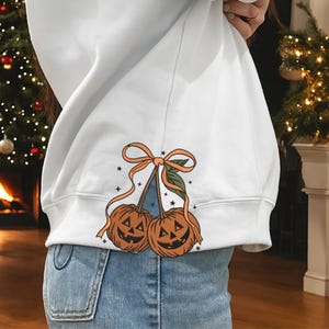 Puede incluir: Sudadera blanca con un diseño de Halloween. El gráfico presenta dos cerezas naranjas con forma de calabaza con caras talladas, conectadas por un tallo y atadas con un lazo naranja. Pequeñas estrellas negras están dispersas alrededor del diseño.