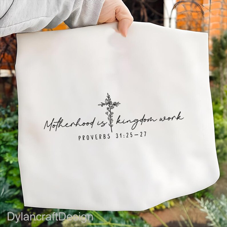 Puede incluir: Tela blanca con el texto "Motherhood is kingdom work" y "PROVERBS 31:25-27" en escritura negra. Un dise&ntilde;o de cruz floral negra est&aacute; centrado encima del texto. La marca "DylancraftDesign" est&aacute; en la parte inferior.