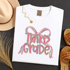 Puede incluir: Camiseta blanca con un diseño bordado colorido que dice "Third Grade" en una fuente juguetona. El diseño incluye elementos de lazo y cinta en tonos de rosa, azul y amarillo. También se ven un collar dorado y un sombrero.