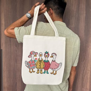 K&ouml;nnte beinhalten: Eine wei&szlig;e Einkaufstasche mit einem Design aus vier G&auml;nsen, die verschiedene Outfits tragen. Die G&auml;nse tragen ein rosa Kleid, einen gelben Regenmantel, ein rotes Erdbeerkleid und ein rosa Kleid mit einer Blumenkrone. Die Tasche hat einen langen Riemen und wird von einer Person gehalten.