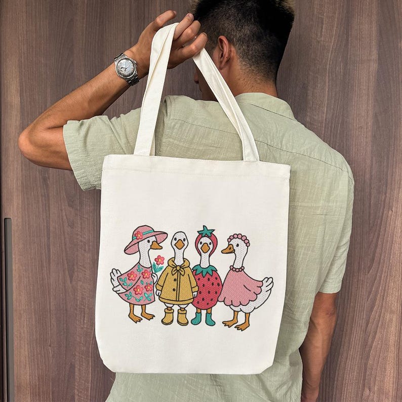 K&ouml;nnte beinhalten: Eine wei&szlig;e Einkaufstasche mit einem Design aus vier G&auml;nsen, die verschiedene Outfits tragen. Die G&auml;nse tragen ein rosa Kleid, einen gelben Regenmantel, ein rotes Erdbeerkleid und ein rosa Kleid mit einer Blumenkrone. Die Tasche hat einen langen Riemen und wird von einer Person gehalten.