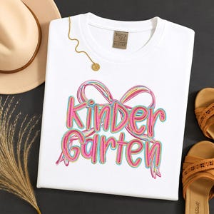 May include: A white t-shirt with the word "Kindergarten" in pink, outlined in blue and yellow, with a matching bow design. A gold necklace and a tan hat are also visible. The shirt is likely for a child.