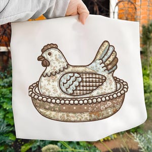 May include: A decorative fabric hen in a nest design. The hen is made of floral patterned fabric in shades of cream, blue, and brown. The nest is a brown bowl shape with a beaded trim. The image is held against a white background.