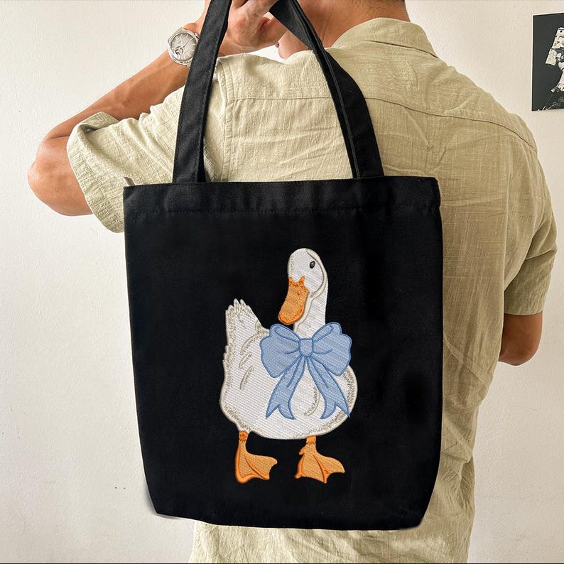 May include: A black canvas tote bag with a white duck embroidered on the front. The duck is wearing a blue bow.