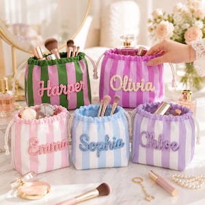 May include: Five small drawstring pouches with vertical stripes in green, pink, purple, and blue. Each pouch features a personalized name in raised, textured lettering: Harper, Olivia, Emma, Sophia, and Chloe. The bags are used for storing cosmetics.
