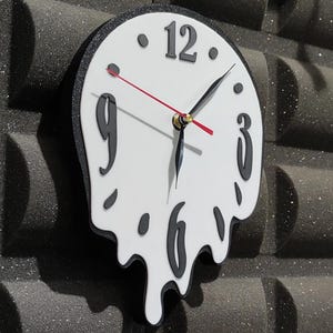 May include: A white wall clock with a melting design. The clock has black numbers and hands, with a red second hand. The clock is mounted on a textured, dark gray wall.
