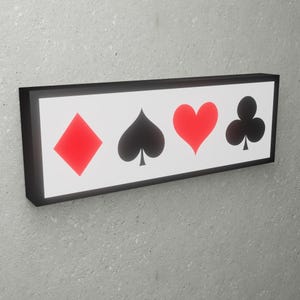 Pode incluir: Uma placa com moldura preta com quatro símbolos de cartas de jogar: um diamante vermelho, um espadilha preto, um coração vermelho e um trevo preto.